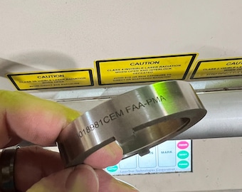 May include: A silver metal ring with the text "018981CEM FAA-PMA" engraved on its surface. The ring is held in a hand, with caution labels visible in the background. The labels warn about laser radiation.