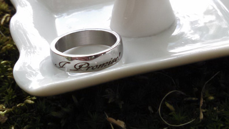 May include: A silver ring with the inscription "I Promise!" is displayed on a white, decorative dish. The ring is set against a backdrop of green moss and a blurred white object.