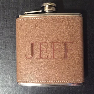 Puede incluir: Un frasco de cuero marr&oacute;n con el nombre "JEFF" grabado en relieve en la parte delantera. El frasco tiene una tapa y un borde plateados, y est&aacute; cosido con hilo blanco alrededor de los bordes. El fondo es oscuro.