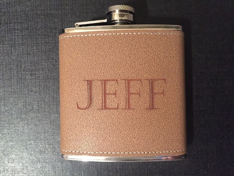Puede incluir: Un frasco de cuero marr&oacute;n con el nombre "JEFF" grabado en relieve en la parte delantera. El frasco tiene una tapa y un borde plateados, y est&aacute; cosido con hilo blanco alrededor de los bordes. El fondo es oscuro.