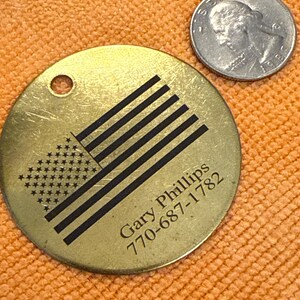 May include: A circular, brass-toned tag featuring a black American flag design, the name "Gary Phillips", and a phone number. A silver coin is next to the tag. The tag has a small hole at the top.