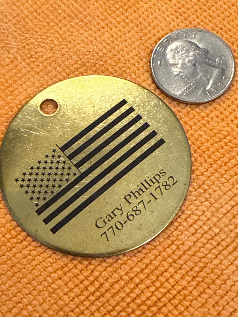 May include: A circular, brass-toned tag featuring a black American flag design, the name "Gary Phillips", and a phone number. A silver coin is next to the tag. The tag has a small hole at the top.