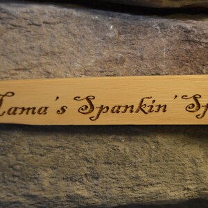 Op de afbeelding: Een lichtgekleurde houten lepel met de tekst "Mama's Spankin' Spoon" in sierlijke letters in het hout ge&euml;tst. De lepel staat tegen een achtergrond van getextureerde, grijze steen.