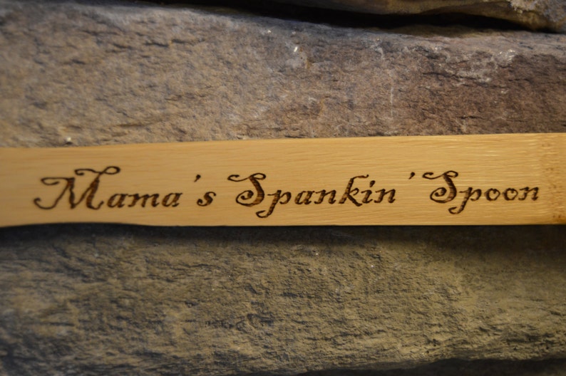 Op de afbeelding: Een lichtgekleurde houten lepel met de tekst "Mama's Spankin' Spoon" in sierlijke letters in het hout ge&euml;tst. De lepel staat tegen een achtergrond van getextureerde, grijze steen.