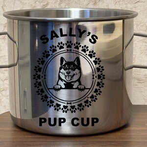 Puede incluir: Un vaso de acero inoxidable con un estampado gráfico negro de un perro centrado dentro de un marco circular de huellas. El texto SALLY'S está impreso sobre el perro y el texto PUP CUP está impreso debajo del diseño en color negro.
