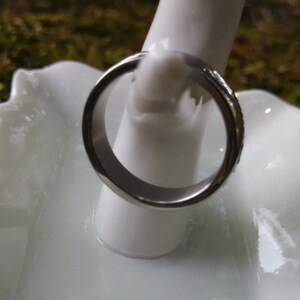 May include: A silver-colored ring with a smooth, polished surface. The ring is displayed on a white, decorative, ceramic dish with a scalloped edge. The background is a blurred view of green moss.