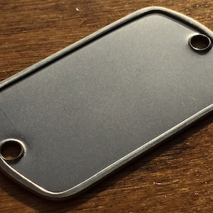 May include: A blank, rectangular dog tag with rounded corners and two circular holes. The tag has a brushed, grey finish and a silver-coloured border. The tag is shown on a wooden surface.