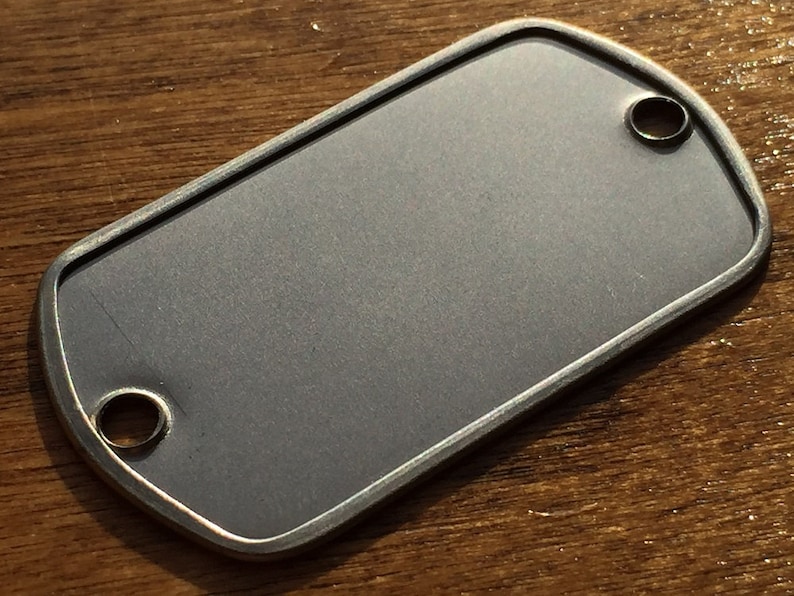 May include: A blank, rectangular dog tag with rounded corners and two circular holes. The tag has a brushed, grey finish and a silver-coloured border. The tag is shown on a wooden surface.