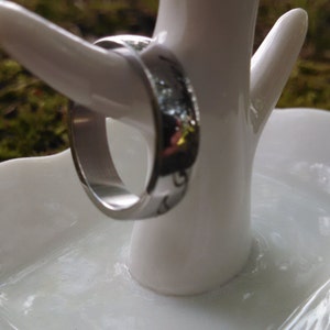 May include: A silver ring is displayed on a white ceramic ring holder. The ring has a polished finish and a subtle design. The holder is shaped like a small tree with a dish base, set against a blurred green background.
