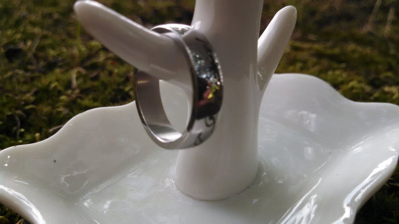 May include: A silver ring is displayed on a white ceramic ring holder. The ring has a polished finish and a subtle design. The holder is shaped like a small tree with a dish base, set against a blurred green background.