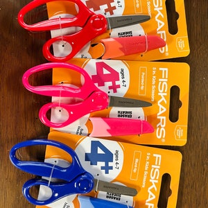 May include: Three pairs of Fiskars 12.7 cm kids scissors in red, pink, and blue, each with a matching eraser sheath. The packaging is orange and white, with the text "ages 4-7" and "5 in. Kids Scissors".