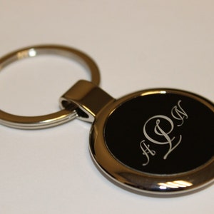 May include: A silver-colored metal keychain with a circular black insert. The insert features the engraved initials "A L N" in a stylized font. The keychain ring is also silver.