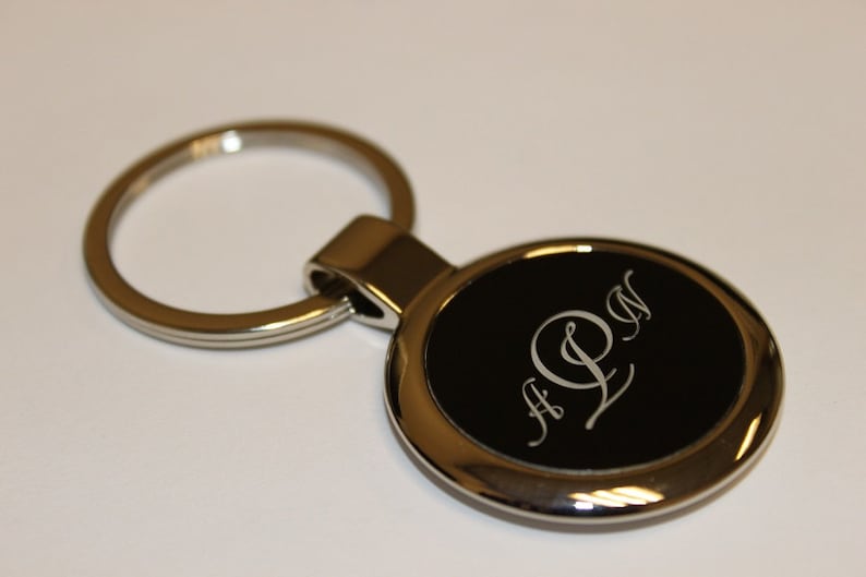 May include: A silver-colored metal keychain with a circular black insert. The insert features the engraved initials "A L N" in a stylized font. The keychain ring is also silver.