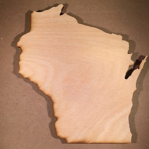 May include: A flat, light-coloured wooden cutout shaped like the state of Wisconsin. The edges of the wood are darkened with a burnt, charred finish, creating a distinct outline that highlights the geographical borders of the state shape.