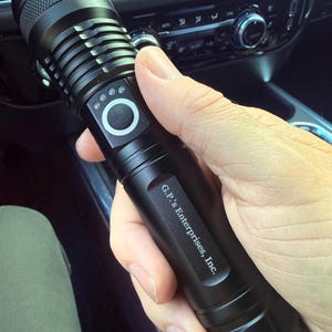 May include: A black cylindrical metal flashlight featuring a textured grip near the head, a circular power button surrounded by four small indicator lights, and the text G.P.'s Enterprises, Inc. printed in white on the side of the handle.