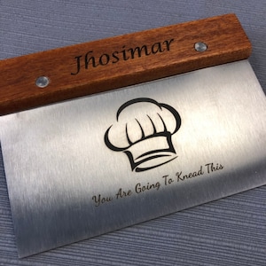 May include: A stainless steel dough scraper with a wooden handle. The handle is engraved with the name "Jhosimar". The scraper features a chef's hat graphic and the text "You Are Going To Knead This".