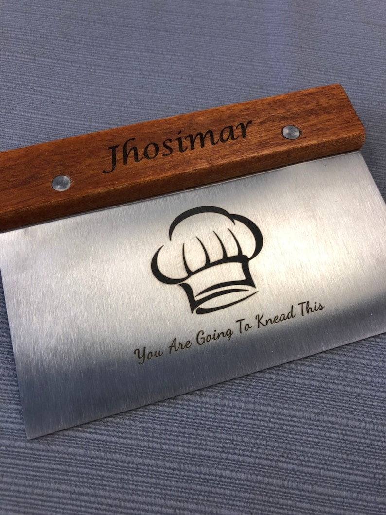 May include: A stainless steel dough scraper with a wooden handle. The handle is engraved with the name "Jhosimar". The scraper features a chef's hat graphic and the text "You Are Going To Knead This".