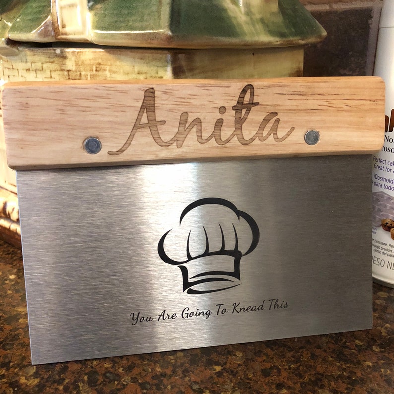 May include: A stainless steel dough scraper with a wooden handle. The handle is engraved with the name "Anita." Below the handle, the scraper features a chef's hat graphic and the text "You Are Going To Knead This."