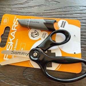 May include: Fiskars brand scissors with black handles and metal blades, featuring the name Sammy printed on the blade. The scissors are packaged on an orange and white cardboard backing that includes a gray eraser sheath and text indicating lefty blades.