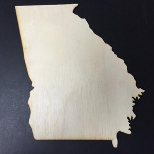 Puede incluir: Un recorte de madera de color claro y plano con la forma del estado de Georgia. La superficie muestra patrones de veta de madera natural y los bordes parecen ligeramente oscurecidos, lo que indica un acabado cortado con láser y uniforme.