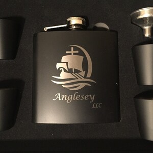 Pu&ograve; includere: Un set di fiaschetta nera con quattro bicchierini abbinati e un imbuto. La fiaschetta presenta un logo di nave argentato inciso e il testo "Anglesey LLC". Il set &egrave; presentato in una scatola nera.