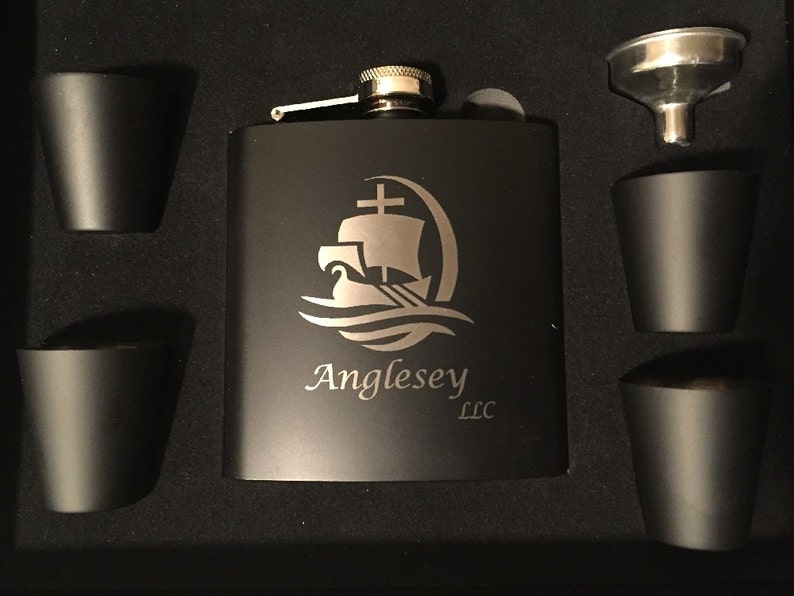 Pu&ograve; includere: Un set di fiaschetta nera con quattro bicchierini abbinati e un imbuto. La fiaschetta presenta un logo di nave argentato inciso e il testo "Anglesey LLC". Il set &egrave; presentato in una scatola nera.