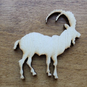 May include: A wooden cutout of a mountain goat, showing a detailed silhouette of the animal. The goat has large curved horns and is standing on four legs. The cutout is light brown and sits on a wooden surface.