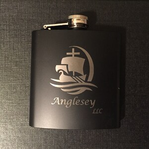 Puede incluir: Petaca negra de acero inoxidable con un dise&ntilde;o plateado de barco y olas. El texto "Anglesey LLC" est&aacute; debajo del dise&ntilde;o. La petaca tiene una tapa con bisagras y una parte superior plateada.
