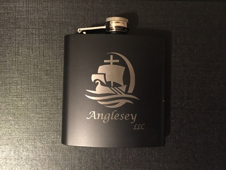 Puede incluir: Petaca negra de acero inoxidable con un dise&ntilde;o plateado de barco y olas. El texto "Anglesey LLC" est&aacute; debajo del dise&ntilde;o. La petaca tiene una tapa con bisagras y una parte superior plateada.