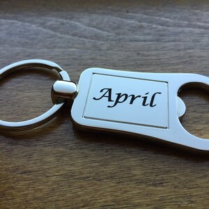 May include: A silver-colored keychain with the month "April" printed in black script. The keychain has a rectangular plate with rounded edges and a bottle opener feature. The background is a wooden surface.