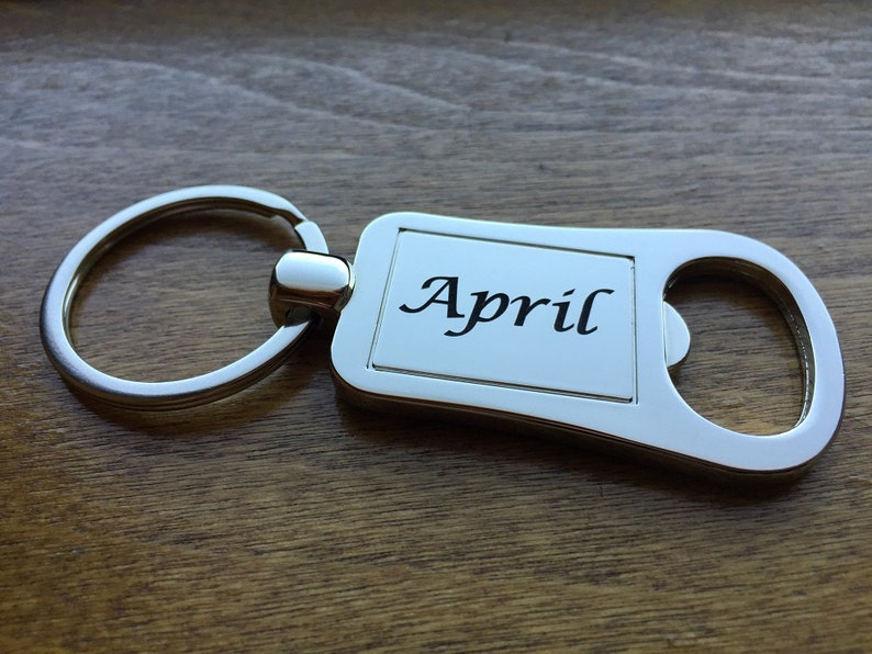 May include: A silver-colored keychain with the month "April" printed in black script. The keychain has a rectangular plate with rounded edges and a bottle opener feature. The background is a wooden surface.