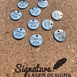 May include: A collection of silver-colored, circular charms, each engraved with "Class of 2024." The charms have a small hole at the top for attaching to a chain or other item. The charms are scattered on a textured, brown surface.