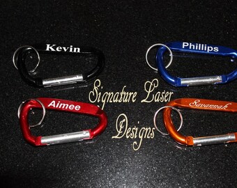 May include: Four carabiner keychains in black, blue, red, and orange, each with a name engraved in white. The keychains are oval-shaped with a spring-loaded gate and a silver key ring. The text "Signature Laser Designs" is also visible.