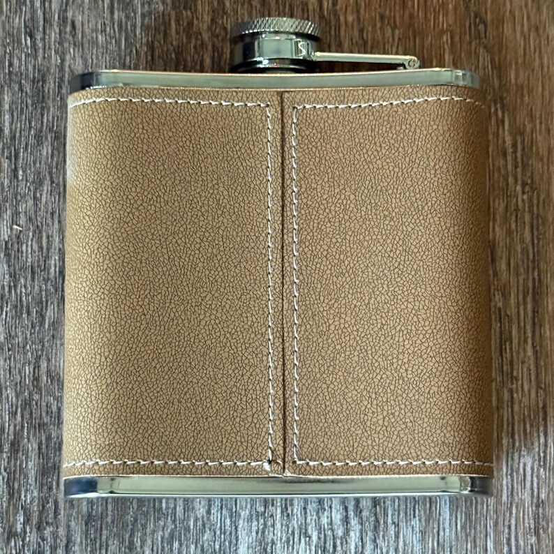 May include: A rectangular metal flask wrapped in textured tan leather with white contrast stitching. The flask features a silver-toned metal frame and a matching silver-toned screw-top cap attached to a hinged arm at the top centre of the container.