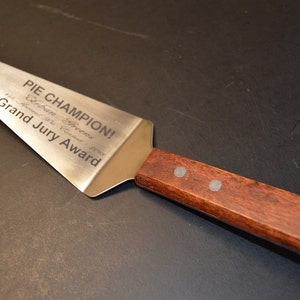 Puede incluir: Una esp&aacute;tula para pastel de acero inoxidable con mango de madera. La esp&aacute;tula est&aacute; grabada con "PIE CHAMPION!" y "Grand Jury Award". El mango es de color marr&oacute;n oscuro y est&aacute; sujeto a la esp&aacute;tula con dos remaches plateados.
