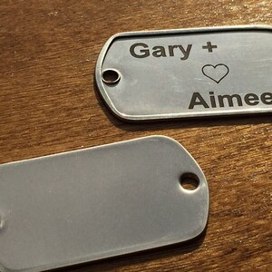 May include: Two silver dog tags. One tag has the names "Gary" and "Aimee" with a heart symbol engraved. Each tag has a hole at one end. The tags are on a wooden surface.