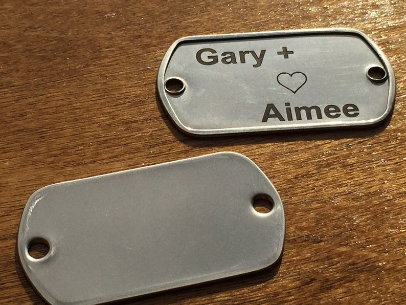 May include: Two silver dog tags. One tag has the names "Gary" and "Aimee" with a heart symbol engraved. Each tag has a hole at one end. The tags are on a wooden surface.