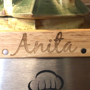 May include: A wooden cutting board with the name "Anita" engraved in a cursive font. The board has a stainless steel blade with a chef's hat logo. The wooden handle is light brown, and the blade is silver.