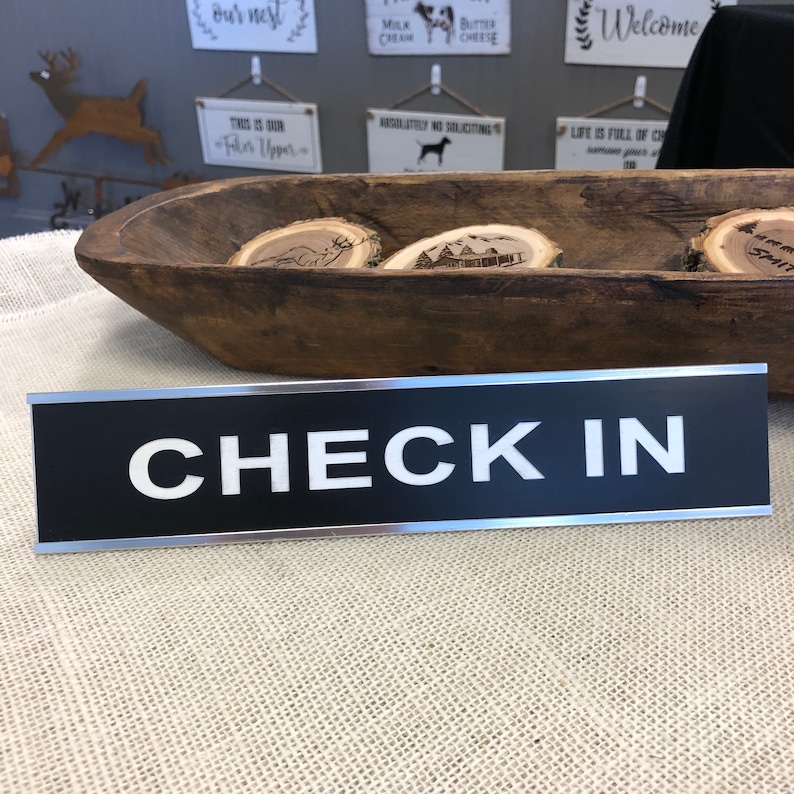 May include: A black and silver "CHECK IN" sign with white lettering. The sign is rectangular and rests on a textured, light-coloured surface. In the background, a wooden bowl holds several wooden slices with etched designs.