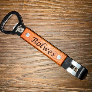 May include: A wooden bottle opener with a stainless steel head. The handle is engraved with the name "Rolwes" and secured with two rivets. The opener is set against a wooden background.