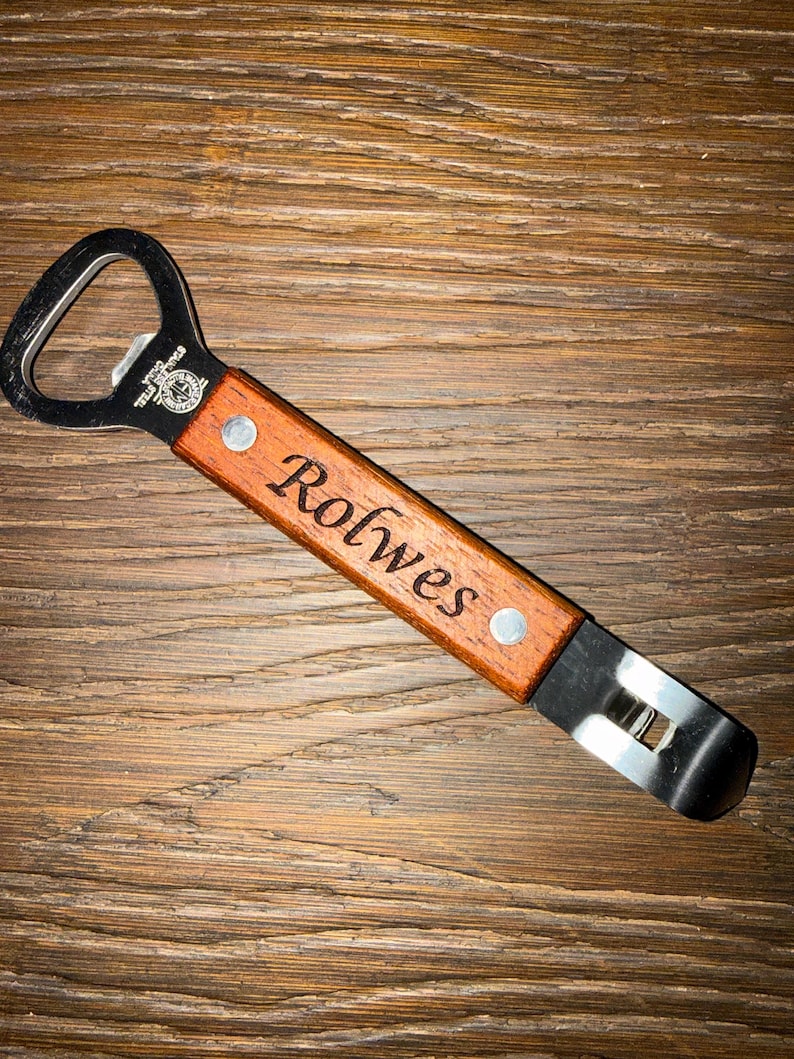 May include: A wooden bottle opener with a stainless steel head. The handle is engraved with the name "Rolwes" and secured with two rivets. The opener is set against a wooden background.