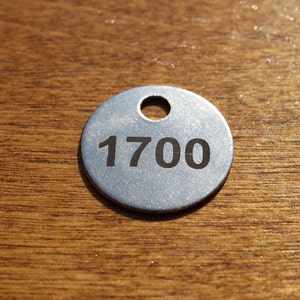 May include: A silver metal tag with the number 1700 printed in black. The tag is round with a hole at the top and is set against a wooden background.