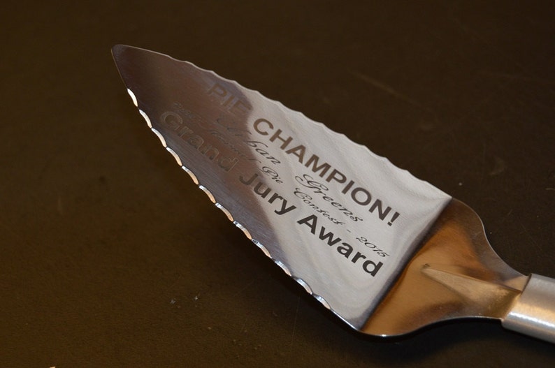 Puede incluir: Una esp&aacute;tula para tartas de acero inoxidable con borde dentado. La esp&aacute;tula est&aacute; grabada con "PIE CHAMPION!" y "Grand Jury Award - 2015". El mango es plateado y la esp&aacute;tula est&aacute; sobre una superficie oscura.
