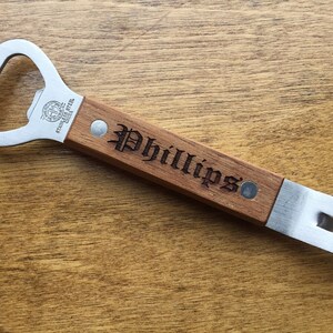 May include: A stainless steel bottle opener with a wooden handle. The handle is engraved with the name "Phillips" in an elegant font. The opener is designed for opening bottles and has a built-in cap lifter.