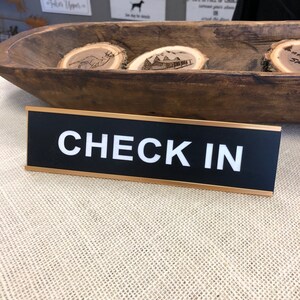 May include: A black sign with the words "CHECK IN" in white. The sign has a gold border and is set on a neutral-coloured surface. In the background is a wooden bowl with decorative wooden slices.