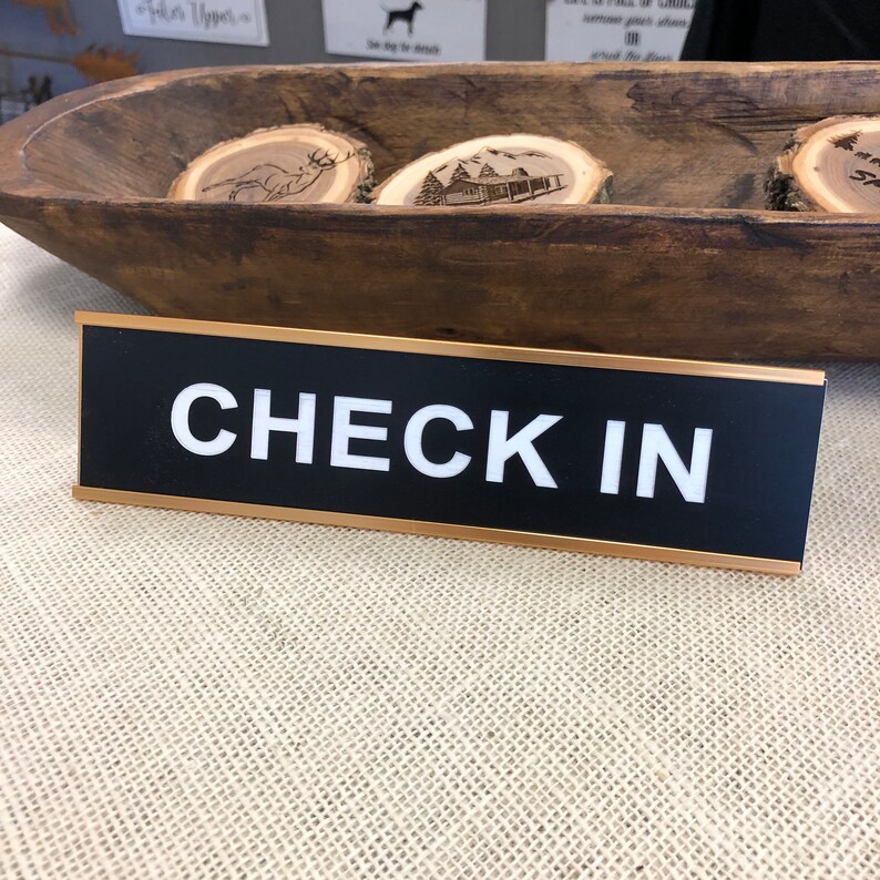 May include: A black sign with the words "CHECK IN" in white. The sign has a gold border and is set on a neutral-coloured surface. In the background is a wooden bowl with decorative wooden slices.