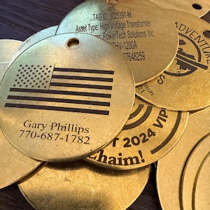 May include: A pile of gold-coloured metal tags. One tag displays an American flag design and the name and phone number "Gary Phillips 770-687-1782". Other tags have text including "TAG ID", "Asset Type", and "2024 VIP".