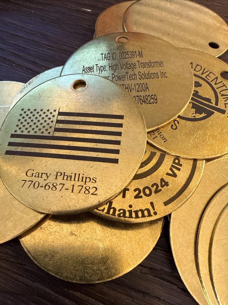 May include: A pile of gold-coloured metal tags. One tag displays an American flag design and the name and phone number "Gary Phillips 770-687-1782". Other tags have text including "TAG ID", "Asset Type", and "2024 VIP".