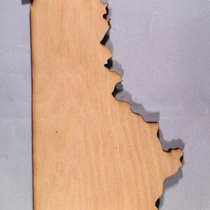 May include: A flat, light brown wooden cutout shaped like the state of Kentucky. The piece features a smooth, natural wood grain surface with dark, charred edges resulting from a laser-cutting process, creating a distinct silhouette of the state.