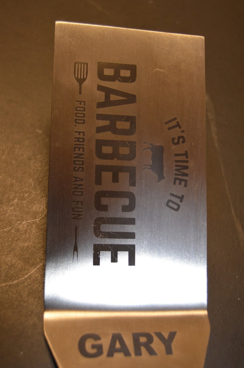 May include: A rectangular stainless steel barbecue spatula featuring the engraved text BARBECUE, IT'S TIME TO, FOOD, FRIENDS AND FUN, and the name GARY. The design includes a small spatula icon, a fork icon, and a cow silhouette in black lettering.